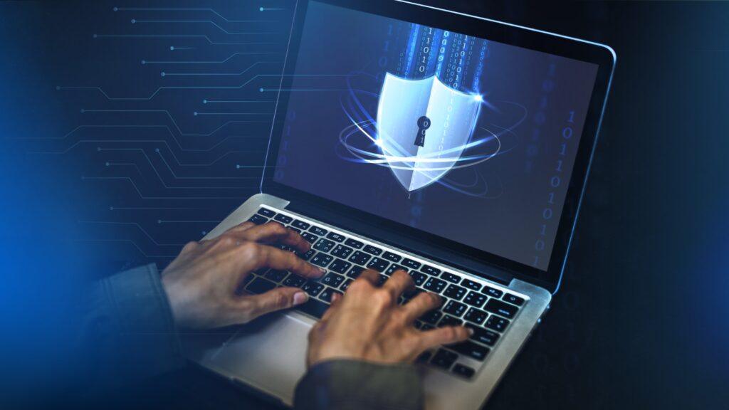 Manos tecleando en un portátil con un icono de escudo y candado en la pantalla, representando ciberseguridad