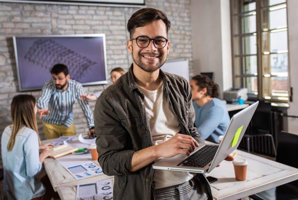 Joven profesional con portátil sonriendo en oficina colaborativa mientras compañeros trabajan en equipo