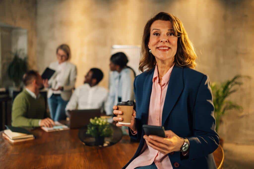 Mujer empresaria sonriente con café y móvil en mano, participando en reunión de trabajo en oficina moderna